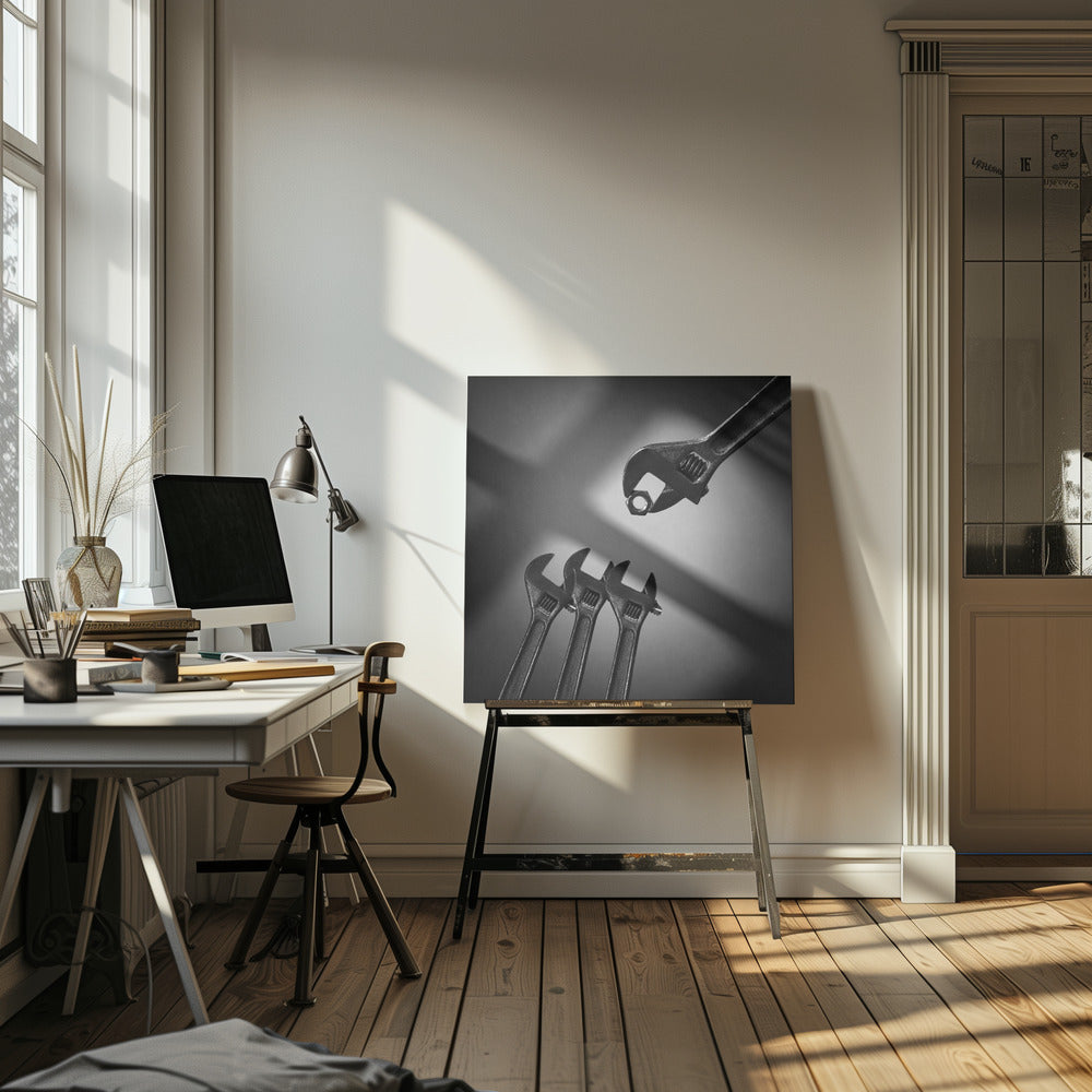 A conceptual black and white photograph in a light wood frame, depicting a large adjustable wrench holding a small nut above three smaller wrenches, which are angled upwards as if they were baby birds waiting to be fed. Print