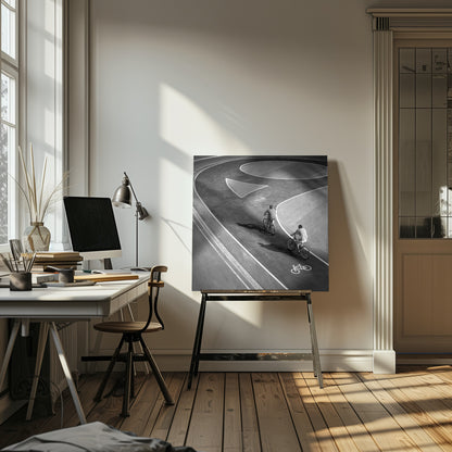 A framed, high-angle black and white photograph of two people cycling on a curving asphalt bike path. The path is marked with bold white lines, including a large arrow and a bicycle symbol, creating a strong graphic pattern. The cyclists cast long shadows on the pavement. Poster