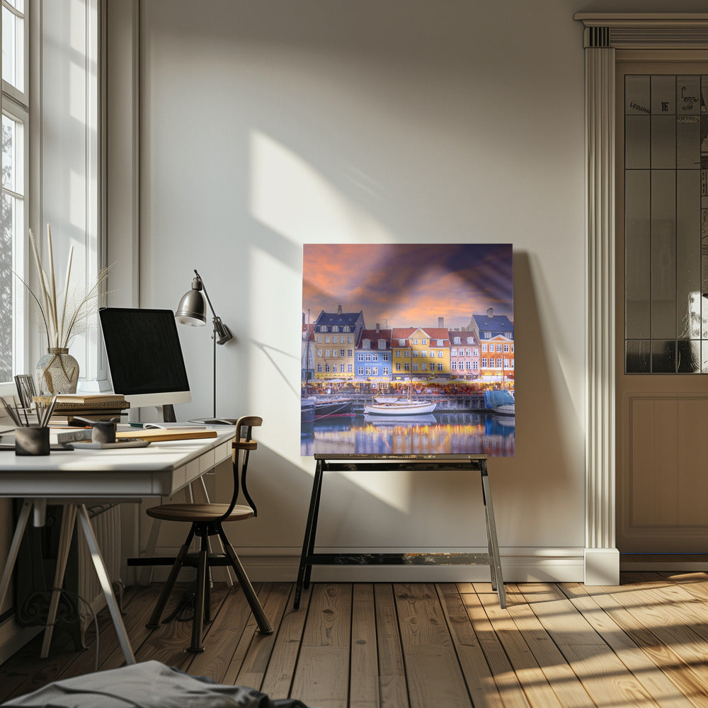 A framed photograph of the Nyhavn harbor in Copenhagen at dusk, featuring a row of brightly colored historic buildings along the canal. Sailboats are moored in the water, which reflects the warm glow of the building lights and the vibrant orange and purple sunset sky. Print