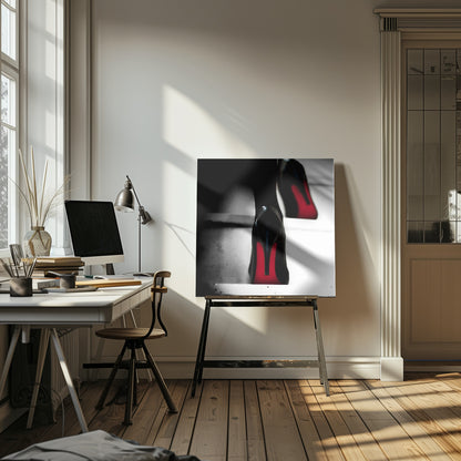 A dramatic, low-angle, black and white photograph focusing on the back of a person wearing glossy black high heels with vibrant red soles, all set within a light wood frame. Artwork