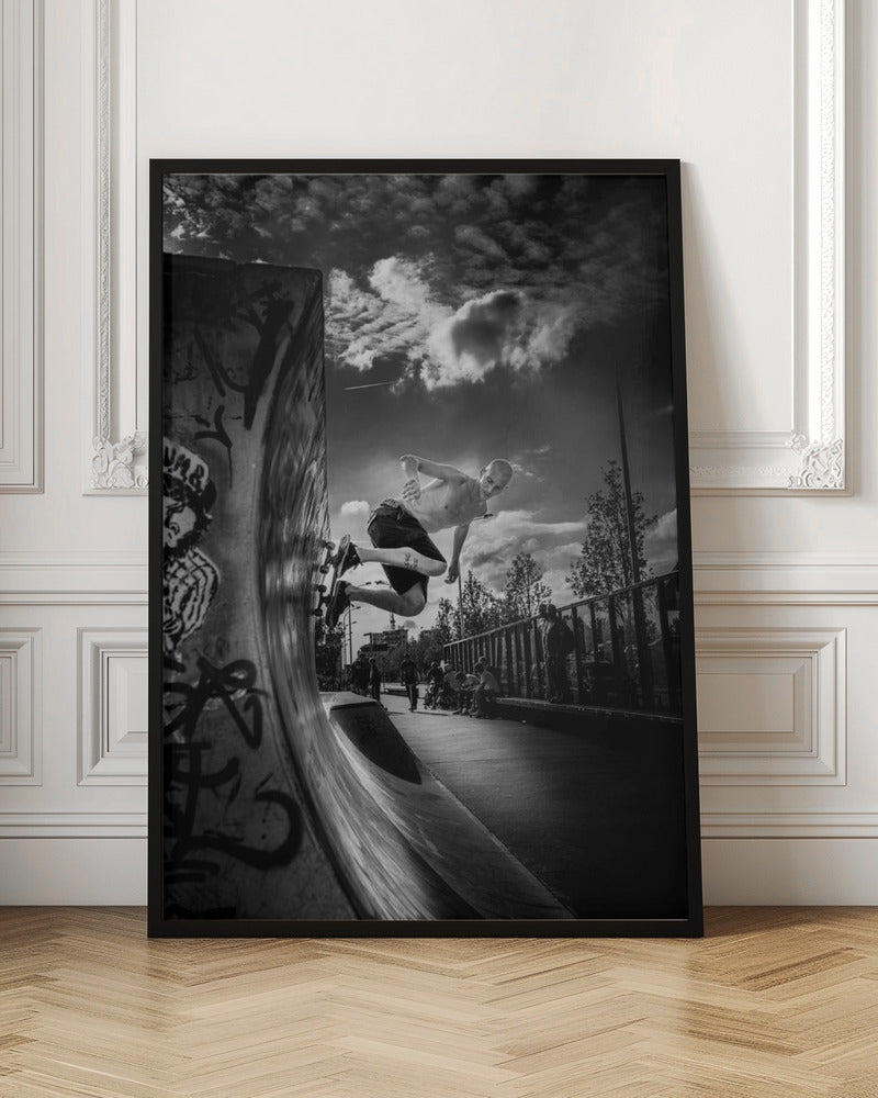 A dramatic black and white, low-angle action shot of a shirtless skateboarder suspended in mid-air at the peak of a graffiti-covered skate ramp, against a backdrop of a cloudy sky. Decor