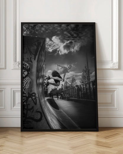 A dramatic black and white, low-angle action shot of a shirtless skateboarder suspended in mid-air at the peak of a graffiti-covered skate ramp, against a backdrop of a cloudy sky. Decor