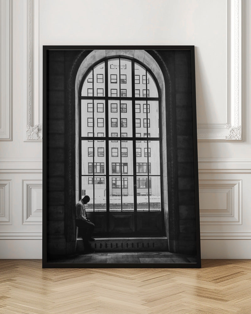 A black and white photograph of a solitary man sitting with his head bowed on the ledge of a massive arched window. The window's dark mullions create a grid over the bright view of another building outside, while the man remains in the shadows, creating a pensive and lonely mood. Poster