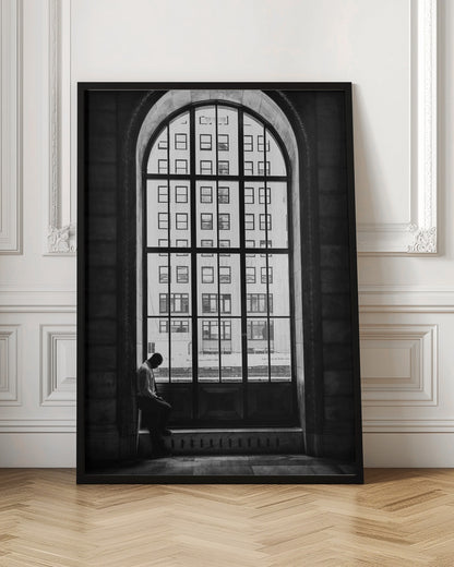 A black and white photograph of a solitary man sitting with his head bowed on the ledge of a massive arched window. The window's dark mullions create a grid over the bright view of another building outside, while the man remains in the shadows, creating a pensive and lonely mood. Poster