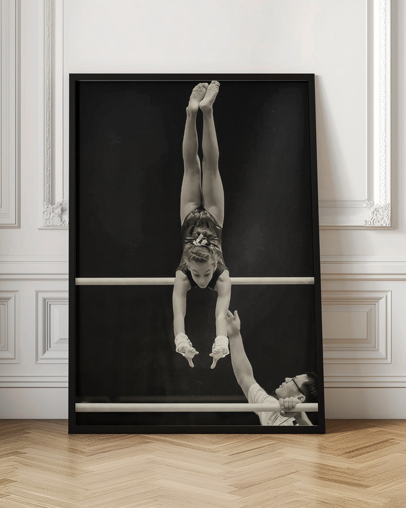 A dramatic black and white photograph of a young female gymnast performing a flawless handstand on the top of two parallel bars. Her body is perfectly vertical against a stark black background. Below, a male coach looks up at her with focused attention, his hand raised nearby, ready to spot her. Decor