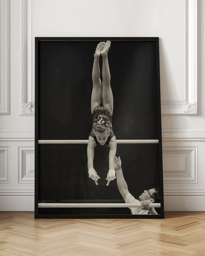 A dramatic black and white photograph of a young female gymnast performing a flawless handstand on the top of two parallel bars. Her body is perfectly vertical against a stark black background. Below, a male coach looks up at her with focused attention, his hand raised nearby, ready to spot her. Decor