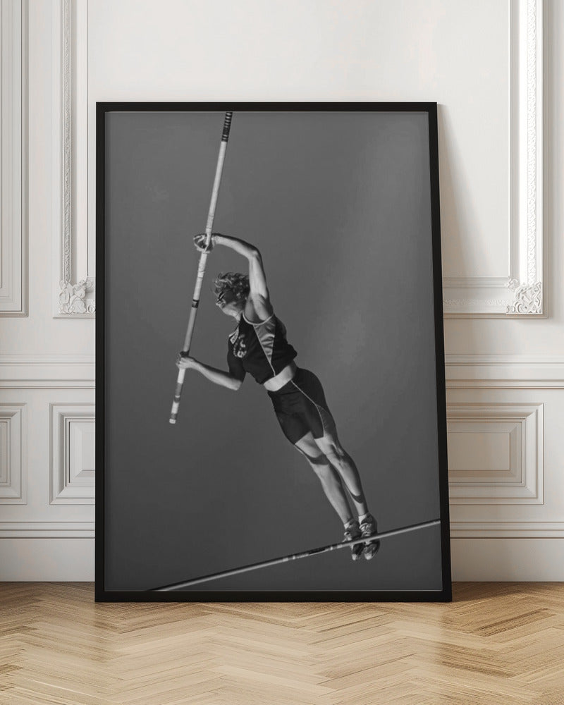 A dynamic black and white, low-angle shot of a female pole vaulter in mid-action. She is suspended in the air, gripping the pole as she prepares to clear the high bar, with her muscular form silhouetted against a plain sky. Print