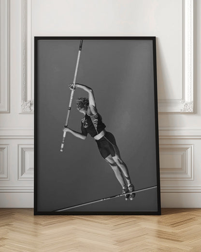 A dynamic black and white, low-angle shot of a female pole vaulter in mid-action. She is suspended in the air, gripping the pole as she prepares to clear the high bar, with her muscular form silhouetted against a plain sky. Print
