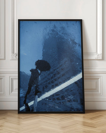 A moody, blue-toned photograph of a reflection in a rainy puddle. A black silhouette of a person holding an umbrella and a distorted brick building are visible, with a single white line crossing the wet street. Poster