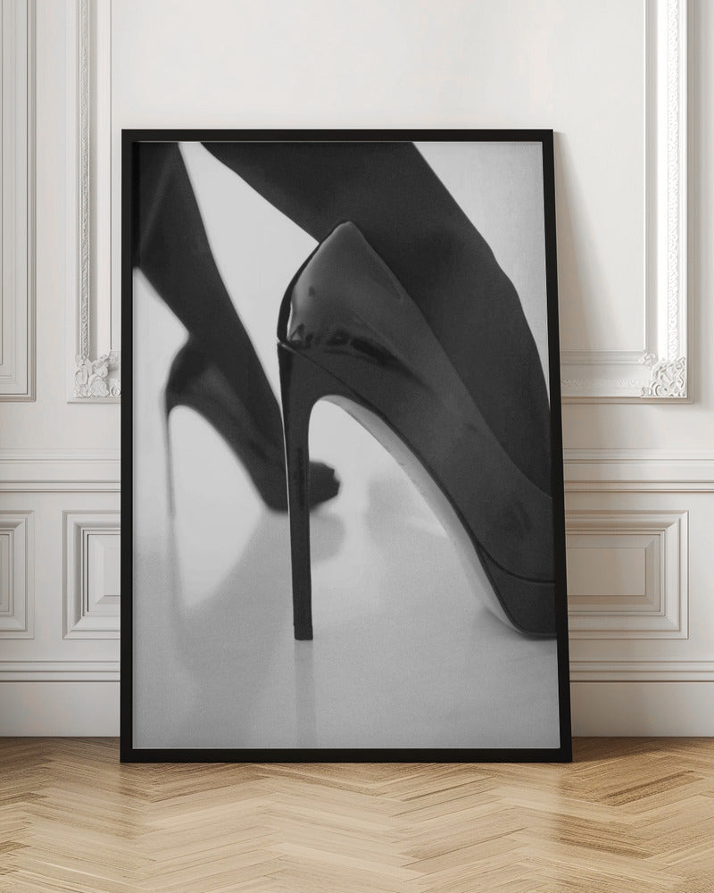 A close-up, black and white photograph of a person wearing shiny black patent leather stilettos and dark stockings. One shoe is in sharp focus in the foreground, highlighting its high arch and thin heel, while the other leg and shoe are softly blurred in the background on a reflective white surface. Decor