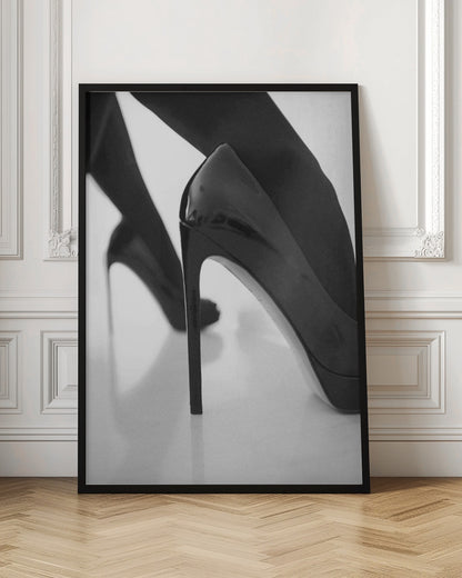A close-up, black and white photograph of a person wearing shiny black patent leather stilettos and dark stockings. One shoe is in sharp focus in the foreground, highlighting its high arch and thin heel, while the other leg and shoe are softly blurred in the background on a reflective white surface. Decor