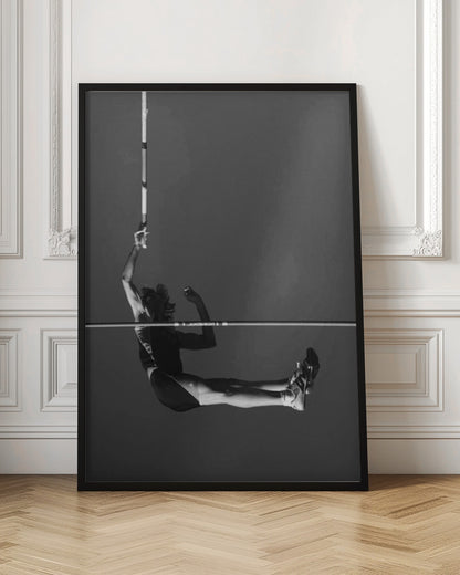 A dramatic black and white photograph of a male pole vaulter in mid-air, successfully clearing the horizontal bar. The athlete's body is parallel to the ground, with one arm still holding the vertical pole, against a solid gray sky. The image is enclosed in a simple black frame. Artwork