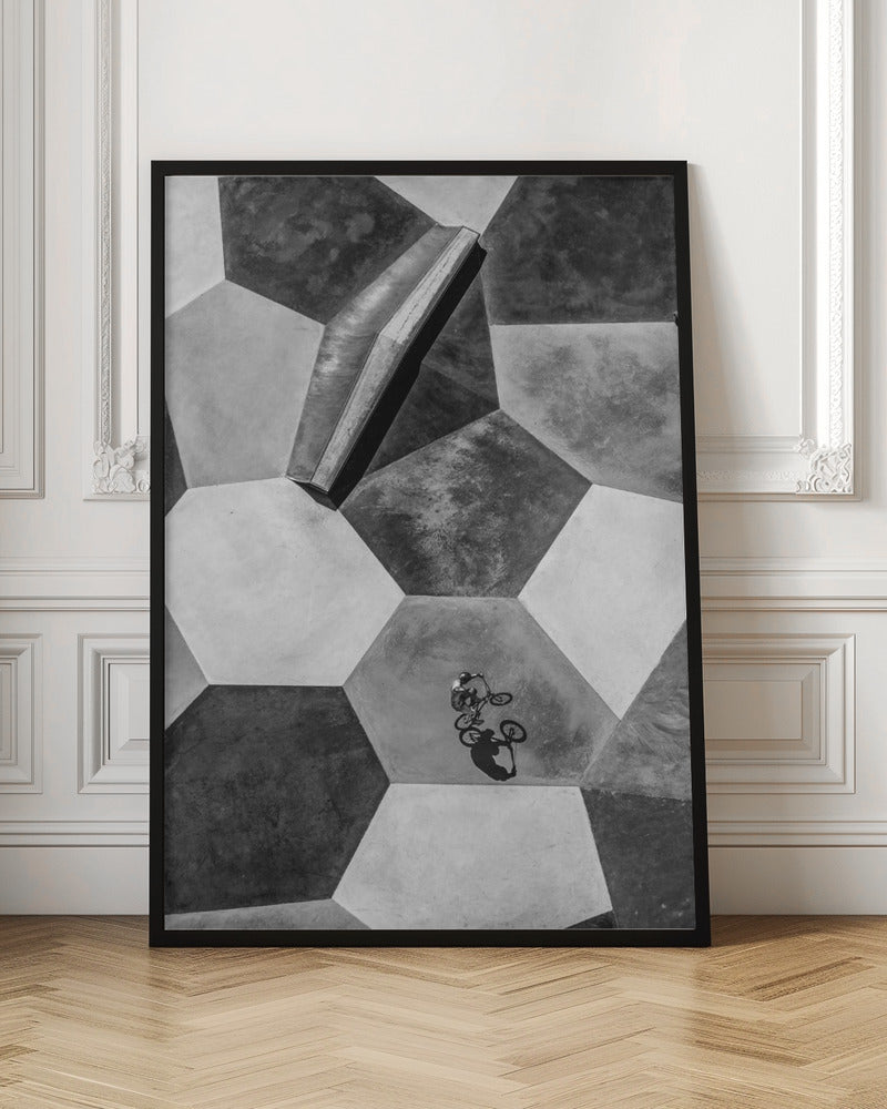 A high-angle, black and white photograph of a BMX rider in a skatepark with a geometric pattern. The ground consists of large, interlocking hexagons in various shades of grey, and the rider is captured mid-action on one of them, with a sharp shadow cast beside them. Artwork