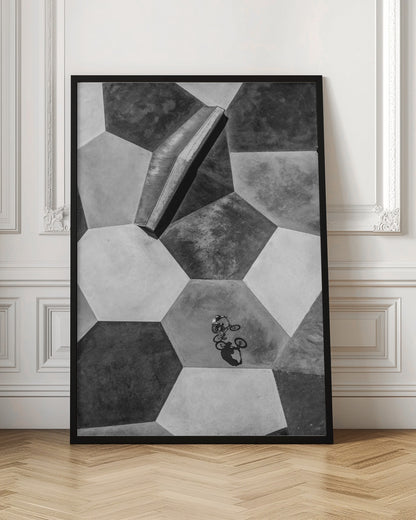 A high-angle, black and white photograph of a BMX rider in a skatepark with a geometric pattern. The ground consists of large, interlocking hexagons in various shades of grey, and the rider is captured mid-action on one of them, with a sharp shadow cast beside them. Artwork