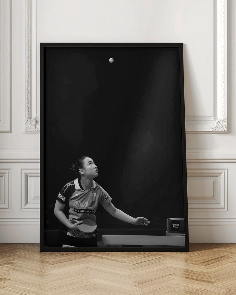 A dramatic black and white vertical photograph of a female table tennis player mid-serve. Positioned in the lower-left, she looks up with intense focus at the white ball suspended high in the air against a solid black background. Poster