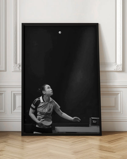 A dramatic black and white vertical photograph of a female table tennis player mid-serve. Positioned in the lower-left, she looks up with intense focus at the white ball suspended high in the air against a solid black background. Poster