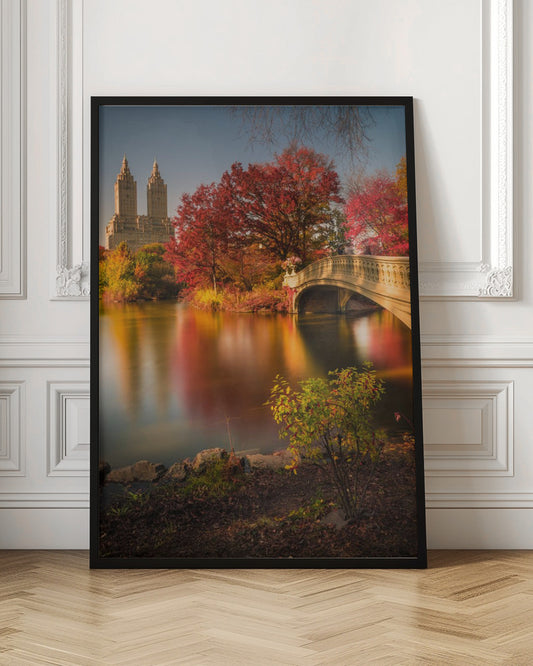 A picturesque autumn scene in Central Park featuring the Bow Bridge over a calm lake reflecting the vibrant red and yellow fall foliage, with the twin towers of The San Remo building visible in the background against a clear blue sky. Decor