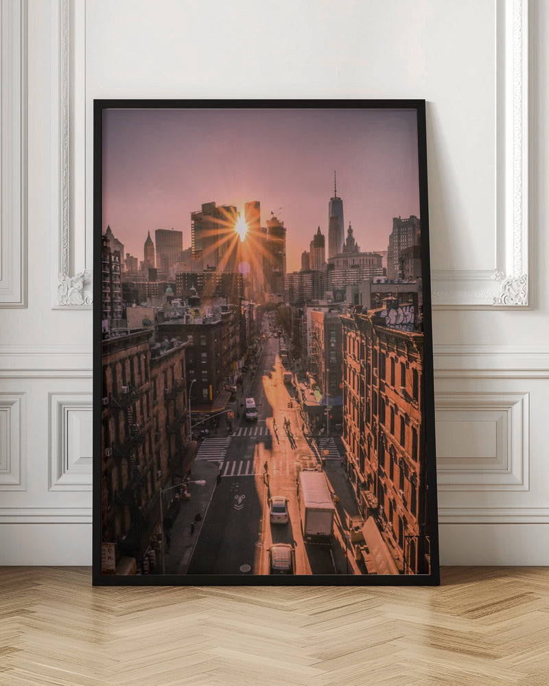 A high-angle view of a New York City street at sunset, with the sun bursting between skyscrapers and casting long golden shadows over the buildings and traffic below. Decor