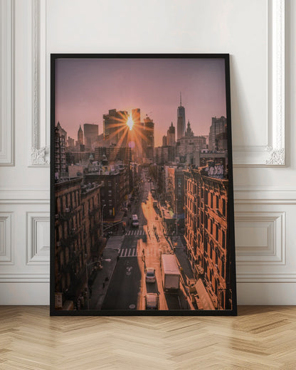 A high-angle view of a New York City street at sunset, with the sun bursting between skyscrapers and casting long golden shadows over the buildings and traffic below. Decor