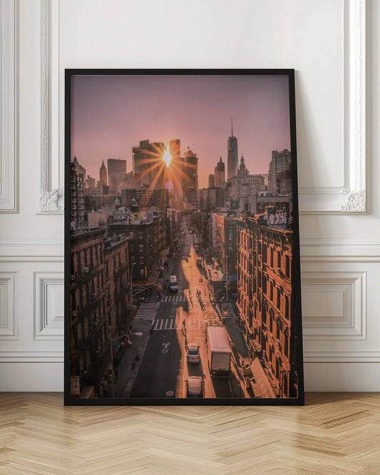 A high-angle view of a New York City street at sunset, with the sun bursting between skyscrapers and casting long golden shadows over the buildings and traffic below. Decor