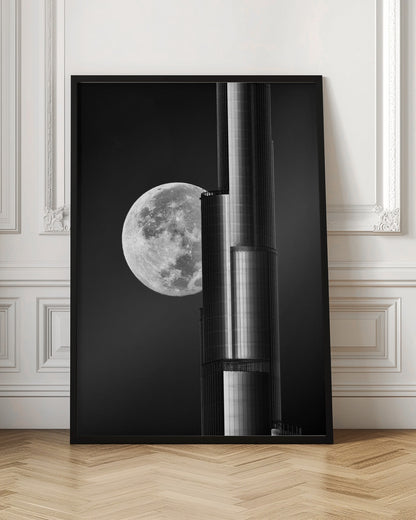 A dramatic black and white photograph of a massive full moon positioned next to a modern, towering skyscraper. The high-contrast image creates a striking juxtaposition between the natural celestial body and the man-made architectural marvel against a dark night sky. Poster