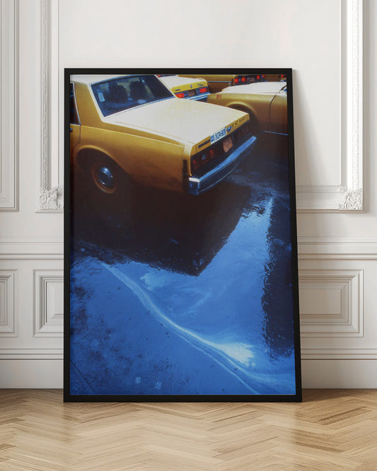 A close-up, angled view of the rear of a yellow taxi cab on a wet street. The car's dark reflection is cast upon a large, vibrant blue puddle in the foreground, with visible ripples from the rain. Wall Art