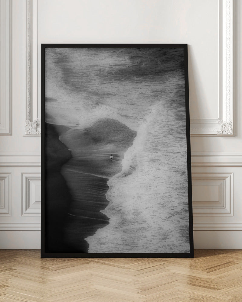 A high-angle, black and white photograph of a lone surfer carrying a surfboard at the water's edge on a dark sand beach. The vast, textured ocean with large, foamy waves crashing ashore dwarfs the solitary figure, creating a sense of scale and solitude. Decor