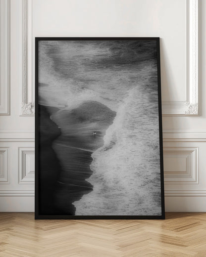 A high-angle, black and white photograph of a lone surfer carrying a surfboard at the water's edge on a dark sand beach. The vast, textured ocean with large, foamy waves crashing ashore dwarfs the solitary figure, creating a sense of scale and solitude. Decor