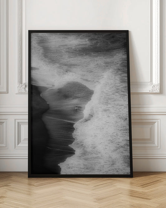 A high-angle, black and white photograph of a lone surfer carrying a surfboard at the water's edge on a dark sand beach. The vast, textured ocean with large, foamy waves crashing ashore dwarfs the solitary figure, creating a sense of scale and solitude. Decor