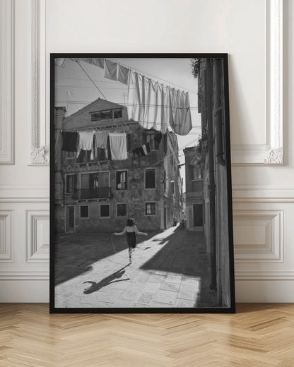 A black and white photograph capturing a girl from behind as she jump-ropes down a sunlit, narrow cobblestone street in Venice. Her long shadow is cast before her, and above, laundry hangs on clotheslines strung between old, weathered European buildings. Wall Art