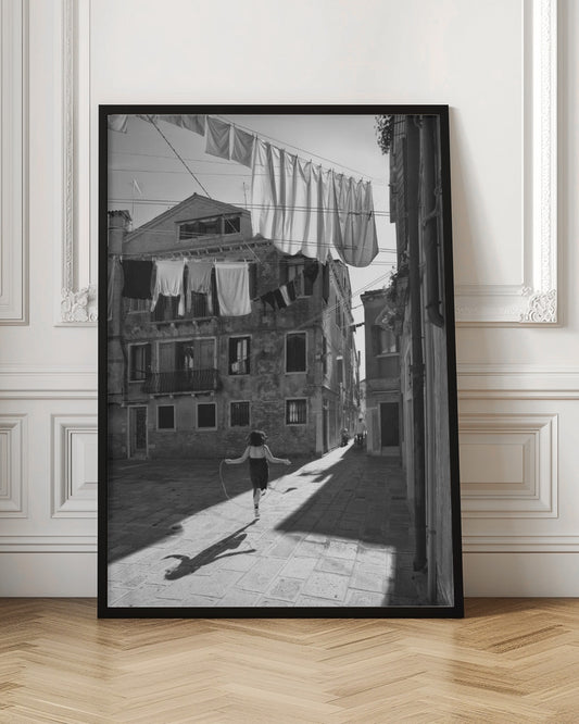 A black and white photograph capturing a girl from behind as she jump-ropes down a sunlit, narrow cobblestone street in Venice. Her long shadow is cast before her, and above, laundry hangs on clotheslines strung between old, weathered European buildings. Wall Art