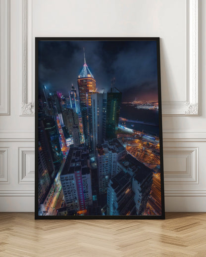 A dramatic high-angle nighttime photograph of a sprawling modern cityscape, with towering skyscrapers illuminated in vibrant blue, orange, and purple neon lights under a dark, cloudy sky. Below, streets are aglow with traffic, and in the distance, a brightly lit harbor meets the dark water. Decor