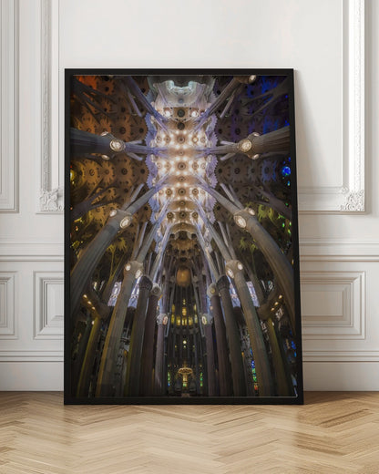A low-angle, symmetrical photograph looking up at the incredibly ornate and complex vaulted ceiling of the Sagrada Familia Basilica. The massive stone columns branch out like trees, supporting the intricately detailed ceiling illuminated by warm, glowing lights and splashes of color from unseen stained glass windows. Poster