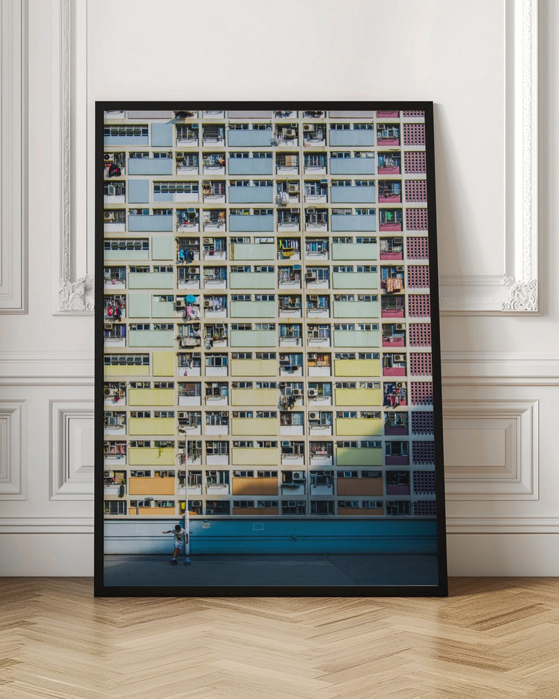 A symmetrical, full-frame shot of a massive apartment building with a repeating grid pattern of windows and balconies. The building is painted in horizontal bands of pastel colors, starting with blue at the top, then green, yellow, and orange. A single boy plays with a soccer ball on the ground level in front of a blue wall. Wall Art