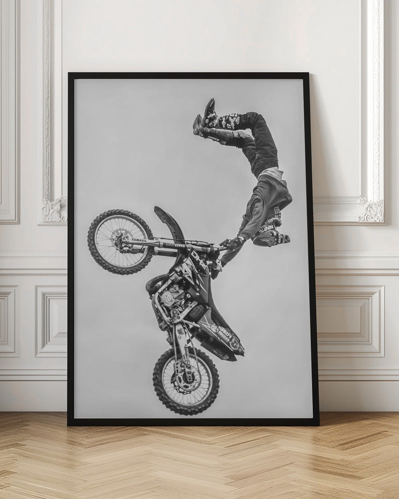 A dynamic black and white action shot of a freestyle motocross rider performing a handstand on the handlebars of their dirt bike mid-air. The bike is nearly vertical against a plain white sky, capturing a moment of extreme athleticism and balance. Poster