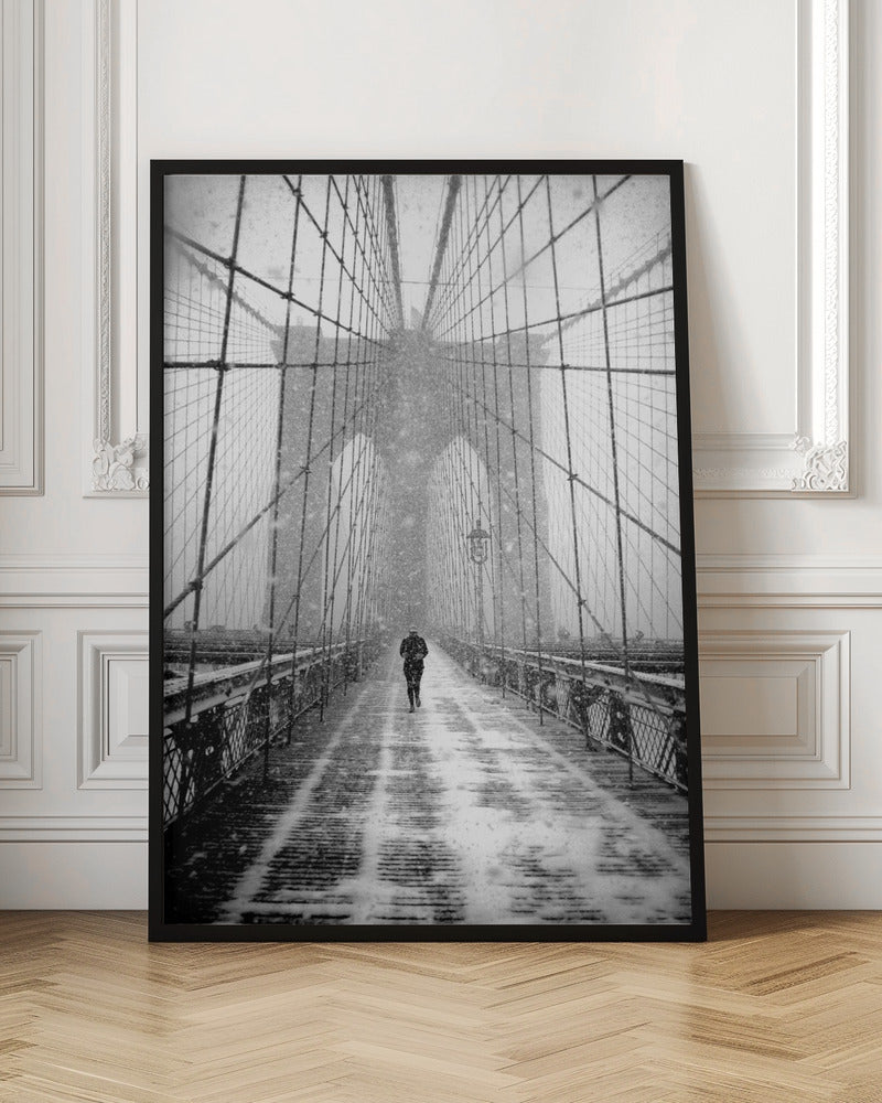 A vertical, black and white photograph of a solitary person walking along the Brooklyn Bridge during a heavy snowstorm. The iconic cables and towers of the bridge create a strong sense of perspective, drawing the eye towards the distant, hazy towers while snow falls all around. Artwork