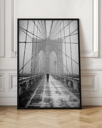 A vertical, black and white photograph of a solitary person walking along the Brooklyn Bridge during a heavy snowstorm. The iconic cables and towers of the bridge create a strong sense of perspective, drawing the eye towards the distant, hazy towers while snow falls all around. Artwork