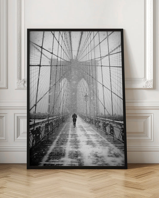 A vertical, black and white photograph of a solitary person walking along the Brooklyn Bridge during a heavy snowstorm. The iconic cables and towers of the bridge create a strong sense of perspective, drawing the eye towards the distant, hazy towers while snow falls all around. Artwork