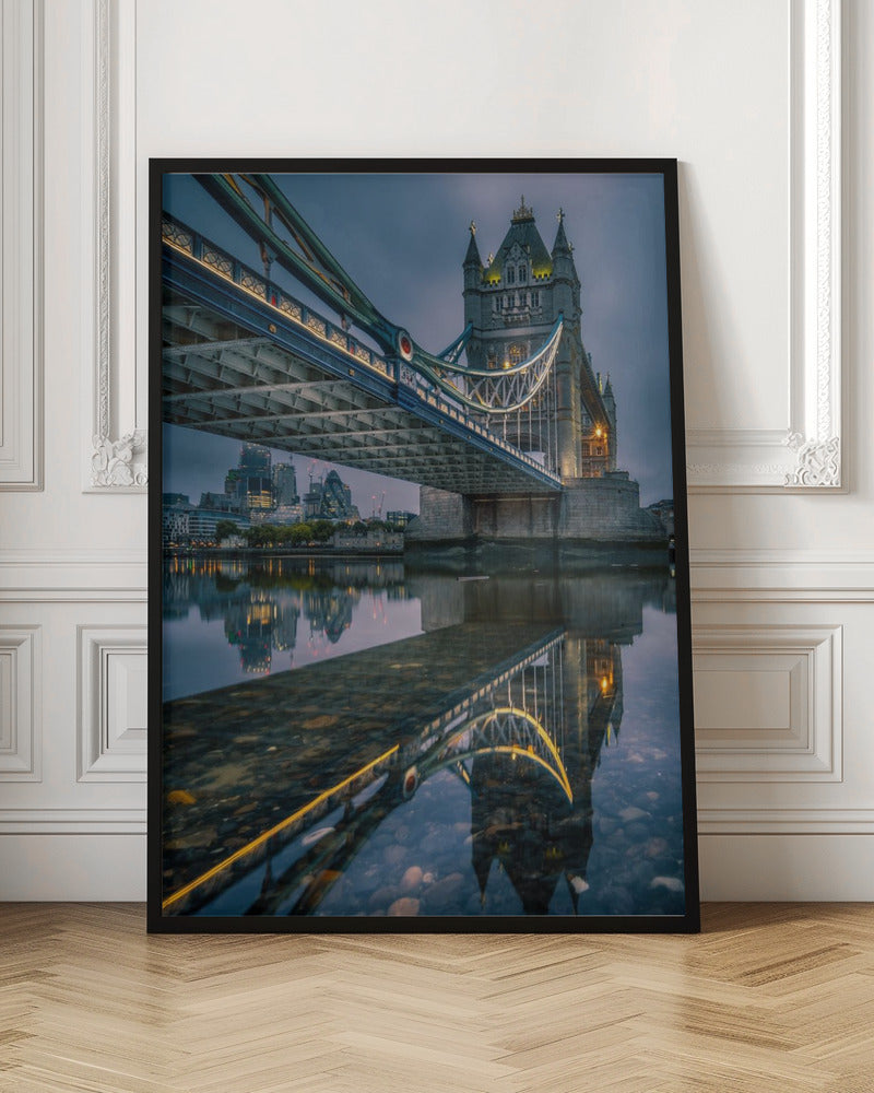 A dramatic, low-angle photograph of London's Tower Bridge at dusk. The iconic bridge is illuminated with warm yellow lights against a dark blue, cloudy sky. The calm River Thames in the foreground creates a stunning, clear reflection of the bridge and the distant city skyline. Print