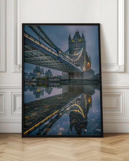 A dramatic, low-angle photograph of London's Tower Bridge at dusk. The iconic bridge is illuminated with warm yellow lights against a dark blue, cloudy sky. The calm River Thames in the foreground creates a stunning, clear reflection of the bridge and the distant city skyline. Print
