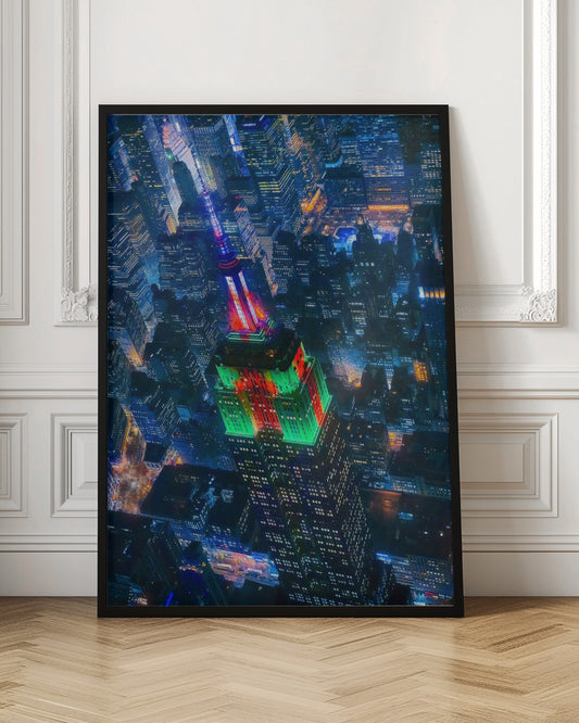 A dramatic high-angle aerial view of the Empire State Building at night, illuminated in festive red and green lights. The surrounding New York City skyline is a dense grid of skyscrapers glowing with blue and white lights, creating a vibrant, sprawling cityscape. Poster