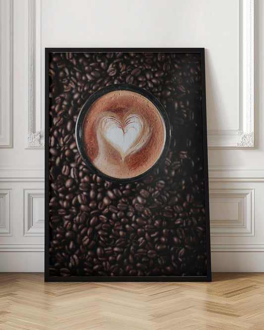 A top-down view of a cappuccino in a black mug with a white heart-shaped foam art design, placed on a bed of dark roasted coffee beans. Artwork