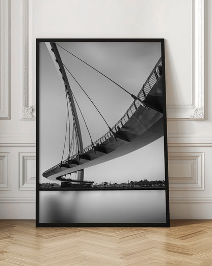A dynamic low-angle, black and white photograph of a modern cable-stayed pedestrian bridge. The bridge curves elegantly over calm, reflective water, with its deck suspended by numerous cables attached to a tall, angled pylon, creating a striking graphic composition against a bright, clear sky. Decor