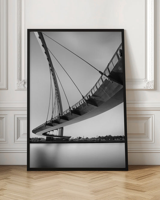 A dynamic low-angle, black and white photograph of a modern cable-stayed pedestrian bridge. The bridge curves elegantly over calm, reflective water, with its deck suspended by numerous cables attached to a tall, angled pylon, creating a striking graphic composition against a bright, clear sky. Decor