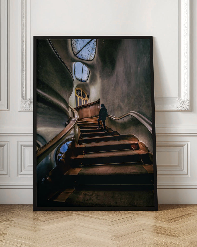 A low-angle view up a dark, winding wooden staircase inside a building with surreal, organic architecture. A person in a dark coat is walking up the stairs towards uniquely shaped windows that show a blue sky, contrasting with the dimly lit, textured gray walls. Print