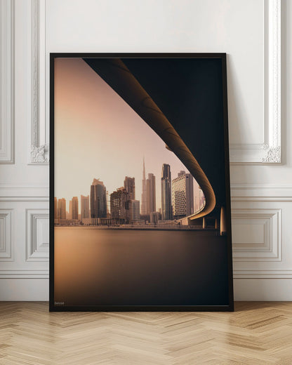 A view of the Dubai skyline, including the Burj Khalifa, framed by the sweeping curve of a bridge in the foreground. The city is bathed in the warm, soft light of a sunrise or sunset, with its reflection visible on the calm water below. Decor