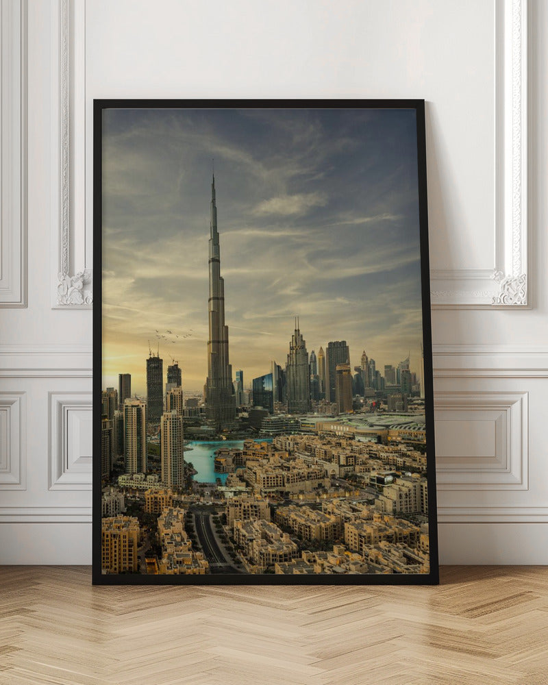 A framed photograph of the Dubai skyline at sunset, with the iconic Burj Khalifa skyscraper dominating the center. Surrounding it are numerous other modern high-rise buildings, a body of turquoise water, and a dense urban landscape in the foreground, all under a cloudy, golden-hued sky. Poster