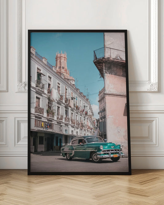 A vintage teal-green classic car sits on a sunny street in a historic city, possibly Havana, Cuba. Weathered apartment buildings with intricate balconies and hanging laundry line the street, with a peach-colored church tower visible in the background against a clear turquoise sky. Poster