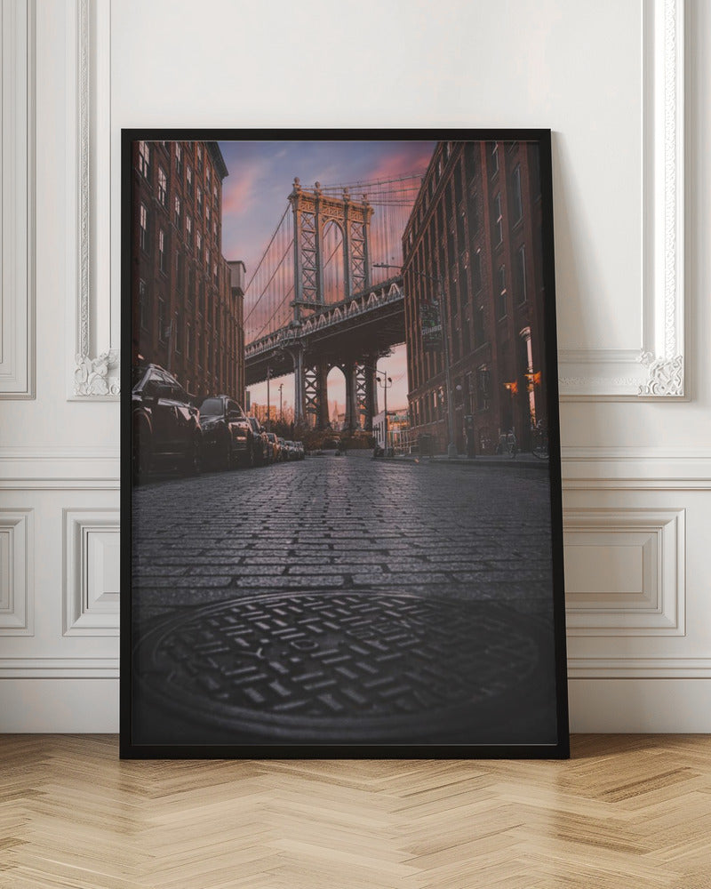 A low-angle photograph taken from a cobblestone street in Dumbo, Brooklyn, looking up towards the Manhattan Bridge at sunset. Red brick buildings frame the view on both sides of the street, with parked cars lining the left. A manhole cover is prominent in the foreground, and the sky is filled with warm pink and orange tones. Print