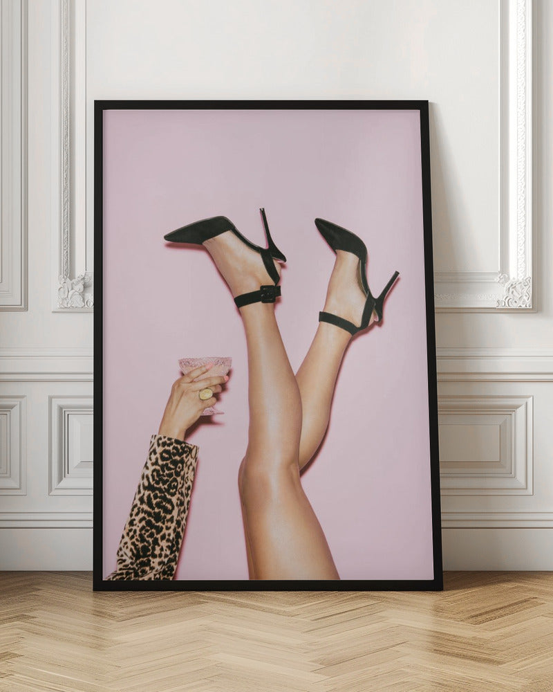 A woman's legs are kicked up in the air against a solid pink background. She is wearing black, pointed-toe, high-heeled shoes with ankle straps. A hand in a leopard print sleeve holds a pink, textured cocktail glass to the side. Artwork